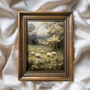 Op de afbeelding: Een ingelijst landschapsschilderij met een pastorale sc&egrave;ne van schapen die in een veld grazen. Het kunstwerk is geplaatst in een goudkleurige lijst en bevat de tekst "YORKSHIRE PRINTS" aan de onderkant. De schildering toont een zonnige dag met bomen en bloemen.