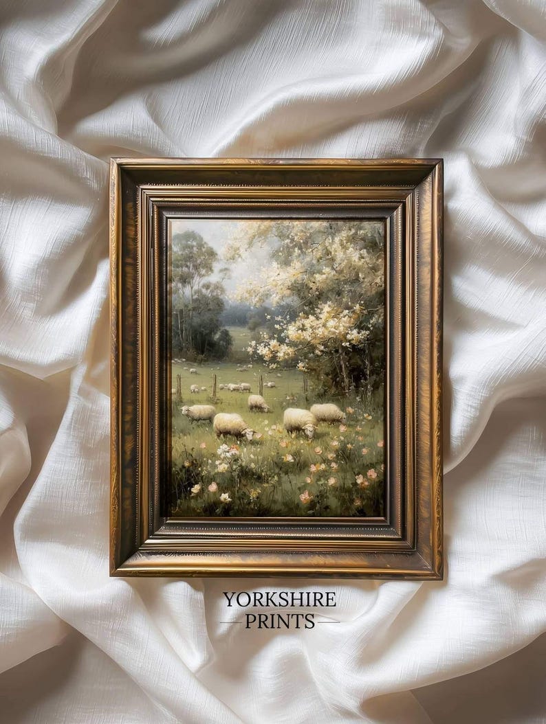 Op de afbeelding: Een ingelijst landschapsschilderij met een pastorale sc&egrave;ne van schapen die in een veld grazen. Het kunstwerk is geplaatst in een goudkleurige lijst en bevat de tekst "YORKSHIRE PRINTS" aan de onderkant. De schildering toont een zonnige dag met bomen en bloemen.