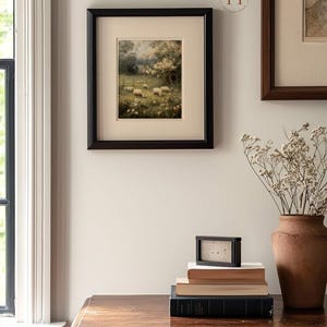 Op de afbeelding: Een ingelijste prent van een landelijke sc&egrave;ne met schapen, geplaatst in een zwarte lijst. Het kunstwerk hangt aan een lichtgekleurde muur, samen met andere ingelijste stukken. Een stapel boeken en een klein zwart frame staan op een houten tafel.