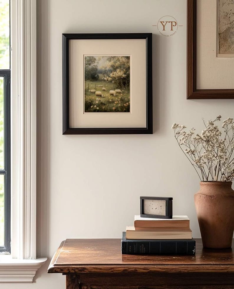 Op de afbeelding: Een ingelijste prent van een landelijke sc&egrave;ne met schapen, geplaatst in een zwarte lijst. Het kunstwerk hangt aan een lichtgekleurde muur, samen met andere ingelijste stukken. Een stapel boeken en een klein zwart frame staan op een houten tafel.