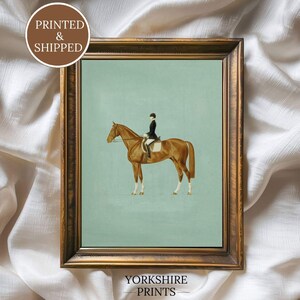 May include: Framed equestrian print featuring a brown horse with a rider in a black coat and helmet, set against a light blue background. The print is in a gold-colored frame. The text "PRINTED & SHIPPED" and "YORKSHIRE PRINTS" are visible.