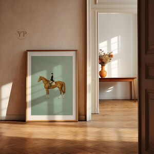 May include: A framed equestrian art print. The artwork features a rider in black attire on a brown horse against a light green background. The frame is a warm, natural wood tone. The print is displayed in a room with hardwood floors.