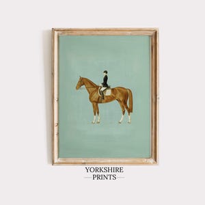May include: A framed equestrian print featuring a brown horse with a rider in a black coat and helmet, set against a light teal background. The artwork is in a wooden frame, with the text "YORKSHIRE PRINTS" at the bottom.
