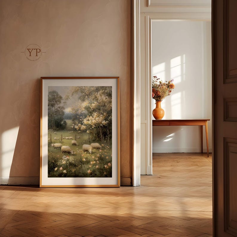 Op de afbeelding: Een ingelijst landschapsschilderij toont schapen die grazen in een veld met bomen en bloemen. Het kunstwerk staat in een gouden lijst en leunt tegen een beige muur. De kamer heeft een houten vloer.