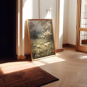 Op de afbeelding: Een ingelijst landschapsschilderij met een pastorale sc&egrave;ne van schapen die grazen in een veld met wilde bloemen. Het kunstwerk heeft een gouden lijst en leunt tegen een muur. De sc&egrave;ne wordt baden in zonlicht, wat een warme sfeer cre&euml;ert.
