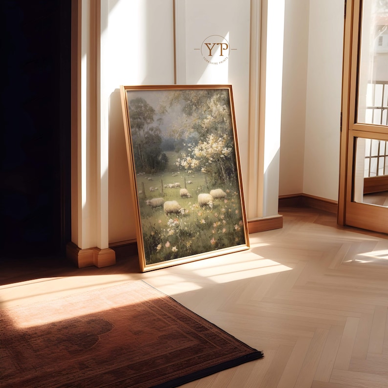 Op de afbeelding: Een ingelijst landschapsschilderij met een pastorale sc&egrave;ne van schapen die grazen in een veld met wilde bloemen. Het kunstwerk heeft een gouden lijst en leunt tegen een muur. De sc&egrave;ne wordt baden in zonlicht, wat een warme sfeer cre&euml;ert.