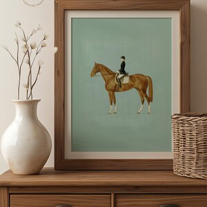 May include: A framed equestrian print featuring a brown horse with a rider in a black coat and helmet. The artwork has a light green background and is displayed in a wooden frame. The print is placed on a wooden dresser with a white vase and a wicker basket.