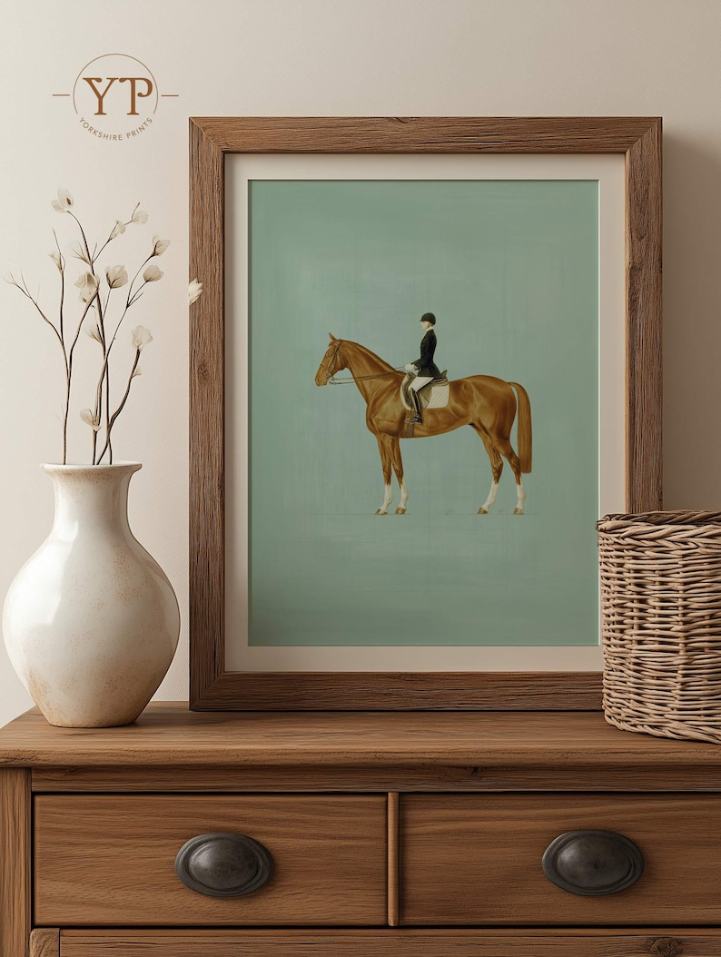 May include: A framed equestrian print featuring a brown horse with a rider in a black coat and helmet. The artwork has a light green background and is displayed in a wooden frame. The print is placed on a wooden dresser with a white vase and a wicker basket.