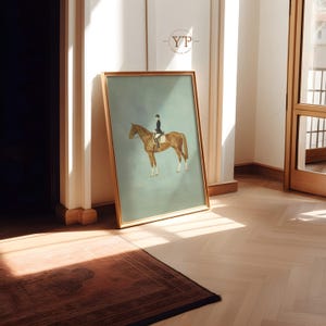 May include: A framed equestrian art print featuring a rider on a brown horse against a muted teal background. The artwork is in a gold frame and leans against a wall in a room with hardwood floors. The print has a vintage aesthetic.