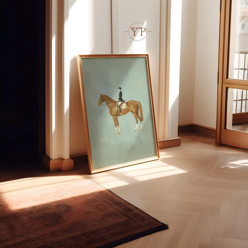 May include: A framed equestrian art print featuring a rider on a brown horse against a muted teal background. The artwork is in a gold frame and leans against a wall in a room with hardwood floors. The print has a vintage aesthetic.