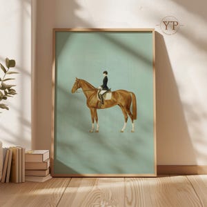 May include: A framed equestrian art print. The artwork features a brown horse with a rider in a black coat and helmet against a light blue background. The horse is in a poised stance, and the print is in a wooden frame.