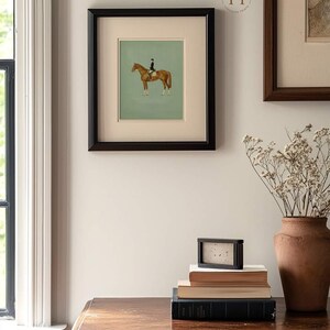 May include: A framed equestrian print featuring a brown horse with a rider in a black coat and hat, set against a light green background. The print is in a black frame, displayed on a wall with other framed art and a wooden table. A stack of books and a vase are on the table.