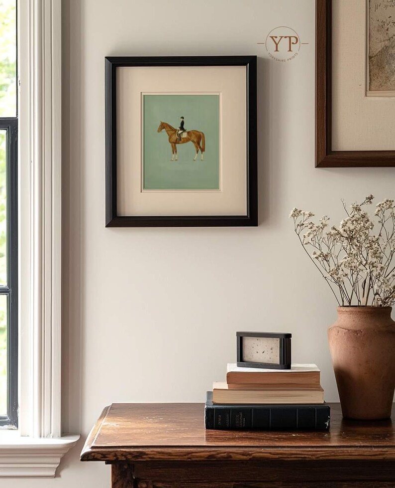 May include: A framed equestrian print featuring a brown horse with a rider in a black coat and hat, set against a light green background. The print is in a black frame, displayed on a wall with other framed art and a wooden table. A stack of books and a vase are on the table.