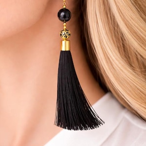 May include: Black tassel earrings with gold-coloured accents. The earrings feature a black bead, a gold filigree bead, and a long black tassel. The earrings are hanging from a gold-coloured hook.