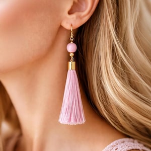 Könnte beinhalten: Rosa Quastenohrringe mit goldfarbenen Akzenten. Die Ohrringe haben eine rosa Perle und eine lange, gefranste Quaste. Die Ohrringe sind mit einem goldfarbenen Haken für durchbohrte Ohren versehen. Ein stilvolles Accessoire.