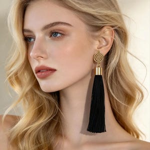 Könnte beinhalten: Ein langer Ohrring mit einem goldfarbenen, kreisförmigen Stecker mit Spiralmuster, der mit einer glatten, goldfarbenen zylindrischen Kappe verbunden ist, an der eine lange, gerade, schwarze Fadenquaste hängt, die vertikal herabfällt.