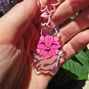 Op de afbeelding: Een roze en bruine bloemenkarakter sleutelhanger met een roze sluiting. Het karakter heeft een lachend gezicht en is omgeven door roze bloemblaadjes. De sleutelhanger wordt in een hand gehouden.