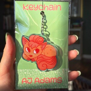 May include: A clear keychain featuring a cartoon fox in shades of orange and red. The keychain is attached to a silver chain and is packaged in a green plastic sleeve. The text "Keychain" and "AJ Adams" are visible.