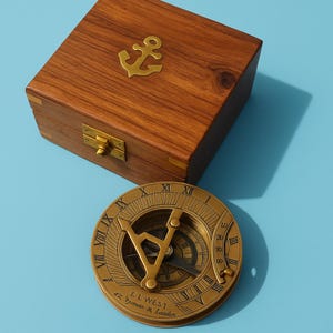 May include: A vintage brass sundial with Roman numerals and a compass, next to a wooden box with a brass anchor emblem. The sundial has the text "EL WEST 42 Jermyn St. London". The box is made of dark wood.