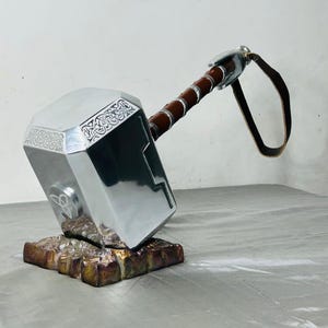 Könnte beinhalten: Ein silberner Hammer mit einem braunen Ledergriff und Riemen. Der Hammerkopf hat aufwendige eingravierte Designs und ruht auf einem strukturierten, braunen Sockel. Der Hammer ist eine Nachbildung von Thors Hammer.