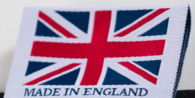 May include: A white fabric label featuring the Union Jack flag in red, white, and blue. The text "MADE IN ENGLAND" is printed in capital letters below the flag. The label has a stitched edge.