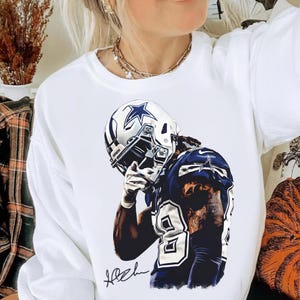 May include: White sweatshirt featuring a graphic of a football player in a Dallas Cowboys uniform. The player is wearing a helmet with the team's star logo and the number 88. The image includes the player's signature.