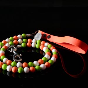 Könnte beinhalten: Eine Hundeleine mit einem Perlendesign in Weiß, Grün und Orange. Die Leine hat einen leuchtend orangefarbenen Griff und einen silberfarbenen Metallverschluss. Die Leine ist auf einer schwarzen Oberfläche aufgerollt.