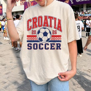 May include: A cream-colored short-sleeve t-shirt featuring a graphic print with the text CROATIA 2026 SOCCER in red and blue. The design includes a central soccer ball illustration and a red and white checkered pattern band across the chest.