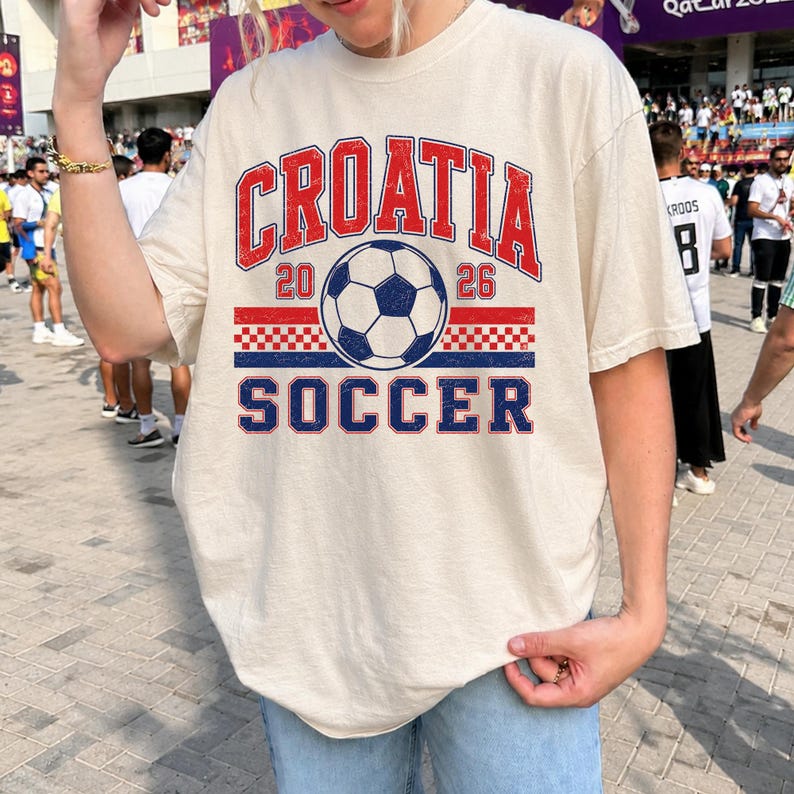 May include: A cream-colored short-sleeve t-shirt featuring a graphic print with the text CROATIA 2026 SOCCER in red and blue. The design includes a central soccer ball illustration and a red and white checkered pattern band across the chest.