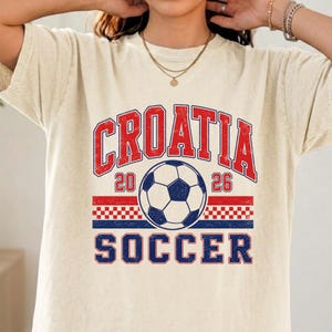 May include: A cream-colored short-sleeve t-shirt featuring a graphic print with the red text CROATIA above a soccer ball, the numbers 20 and 26 on either side, a red and white checkered pattern, and the blue text SOCCER at the bottom.