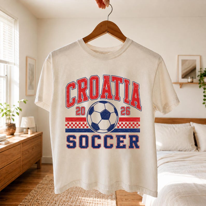 May include: A cream-colored short-sleeve t-shirt featuring a distressed graphic print. The design includes the red text "CROATIA" above a soccer ball, the numbers "20" and "26", a red and blue checkered stripe, and the blue text "SOCCER" below.
