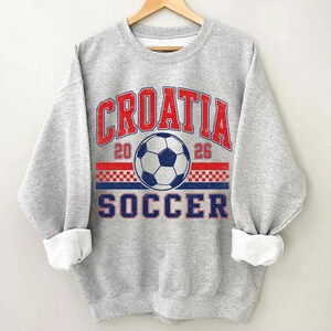May include: A heather gray crewneck sweatshirt featuring a distressed graphic print. The design includes the red text CROATIA, the blue text SOCCER, the numbers 20 and 26, a central soccer ball, and a red and white checkered pattern stripe.