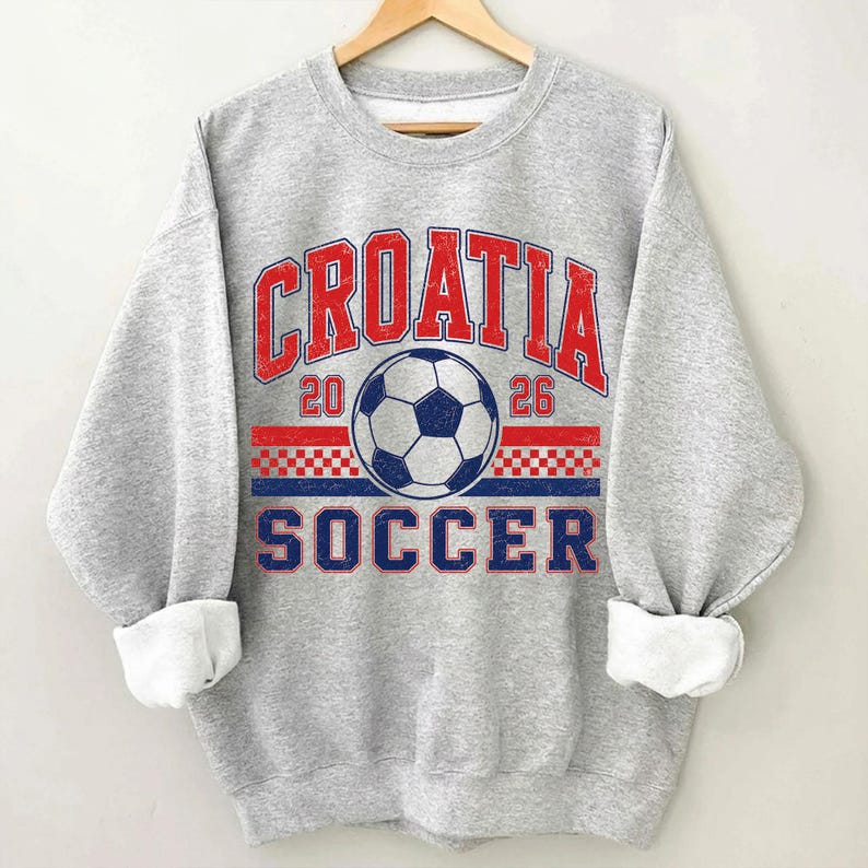 May include: A heather gray crewneck sweatshirt featuring a distressed graphic print. The design includes the red text CROATIA, the blue text SOCCER, the numbers 20 and 26, a central soccer ball, and a red and white checkered pattern stripe.