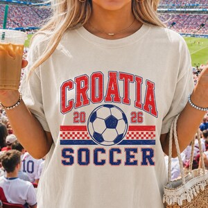 May include: A beige short-sleeve t-shirt featuring a graphic print with the text CROATIA in red, the year 2026, and the word SOCCER in blue. The design includes a central soccer ball illustration and a red and white checkered pattern stripe.