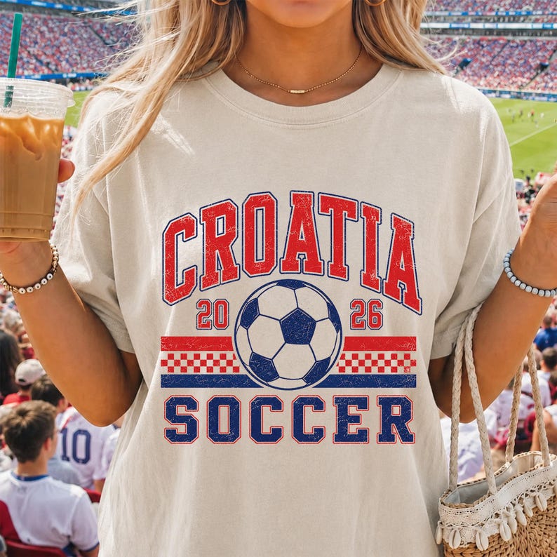 May include: A beige short-sleeve t-shirt featuring a graphic print with the text CROATIA in red, the year 2026, and the word SOCCER in blue. The design includes a central soccer ball illustration and a red and white checkered pattern stripe.
