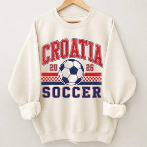 May include: A cream-colored crewneck sweatshirt featuring a distressed graphic print. The design includes the red text "CROATIA" above a soccer ball, the numbers "20" and "26", a red and white checkered pattern, and the blue text "SOCCER".