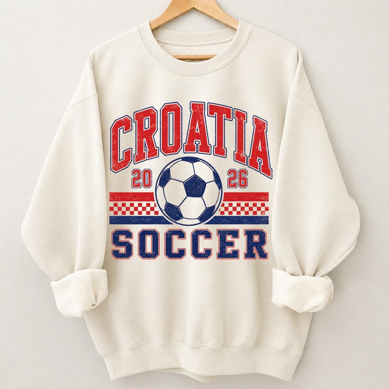 May include: A cream-colored crewneck sweatshirt featuring a distressed graphic print. The design includes the red text "CROATIA" above a soccer ball, the numbers "20" and "26", a red and white checkered pattern, and the blue text "SOCCER".