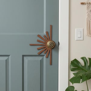 Könnte beinhalten: Eine türkisblaue Tür mit einem türförmigen Türklopfer in Form eines Sonnenstrahls. Die Tür ist von Topfpflanzen umgeben, darunter eine große Monstera-Pflanze. Auf dem Boden liegt ein gemusterter Teppich. Ein Makramee-Wandbehang hängt an der Wand.