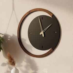 Puede incluir: Un reloj de pared moderno con esfera gris oscuro y manecillas doradas. El reloj está enmarcado en una media luna de madera marrón. Está montado en una pared beige claro, con un diseño minimalista.