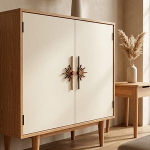 May include: A rectangular wooden cabinet with light oak sides and legs, featuring two off-white doors. The doors are equipped with dark brown, sunburst-shaped vertical handles that meet in the center, creating a distinct decorative focal point.