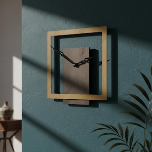 May include: A contemporary square wall clock with a gold-coloured frame and black clock hands. The clock face is a rectangular shape in a darker tone, mounted on a teal wall. The clock is approximately 30 cm square.