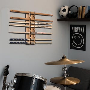 Pode incluir: Um suporte de madeira para baquetas montado numa parede cinza, exibindo vários pares de baquetas. O suporte é feito de madeira castanha clara e tem um design decorativo. Algumas baquetas têm texto. Um poster do Nirvana é visível.