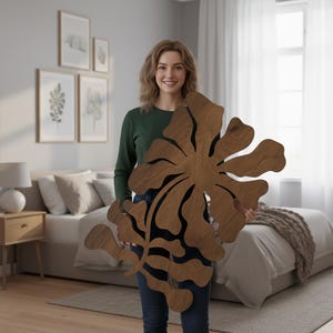 Peut inclure: Grande d&eacute;coration murale florale en bois avec une finition en grain de bois naturel. L'&oelig;uvre d'art pr&eacute;sente un motif de fleur abstrait avec de multiples p&eacute;tales et une tige centrale. La pi&egrave;ce est tenue par une personne dans une pi&egrave;ce &agrave; la d&eacute;coration neutre.