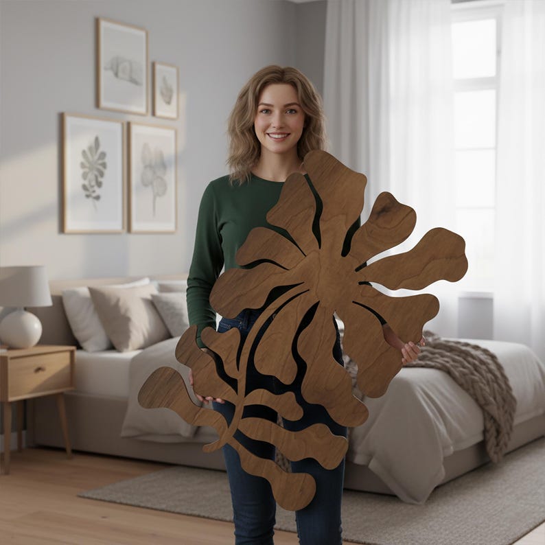 Peut inclure: Grande d&eacute;coration murale florale en bois avec une finition en grain de bois naturel. L'&oelig;uvre d'art pr&eacute;sente un motif de fleur abstrait avec de multiples p&eacute;tales et une tige centrale. La pi&egrave;ce est tenue par une personne dans une pi&egrave;ce &agrave; la d&eacute;coration neutre.