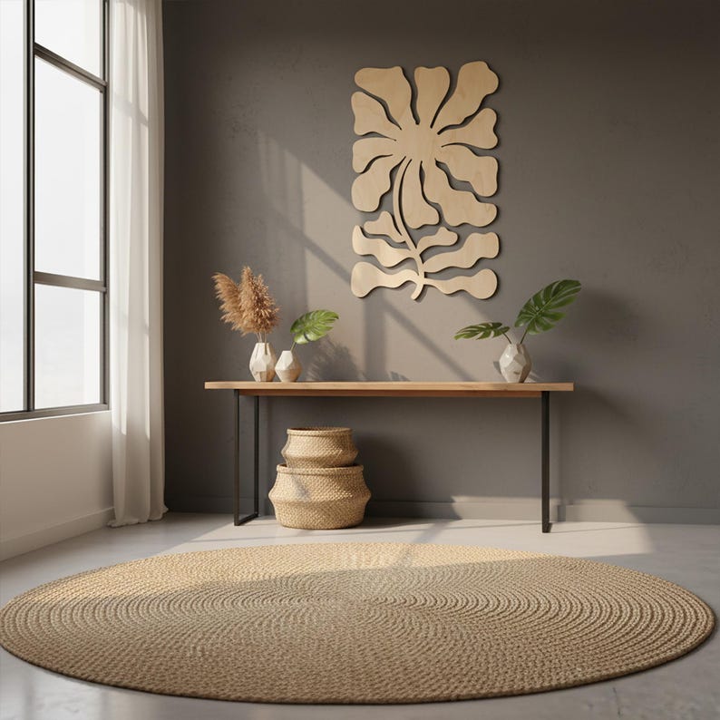 Peut inclure: Un salon aux tons neutres avec un grand tapis rond tiss&eacute;. Une console en bois avec des pieds en m&eacute;tal noir supporte des vases et des plantes. Une grande &oelig;uvre d'art murale en forme de fleur en bois est accroch&eacute;e au mur gris, et deux paniers tress&eacute;s sont empil&eacute;s en dessous.