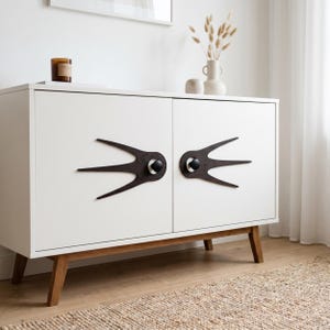 May include: A white sideboard with two doors featuring dark brown, swallow-shaped wooden decorative plates behind round black knobs. The cabinet is supported by four angled wooden legs and has a clean, rectangular frame with a smooth finish.