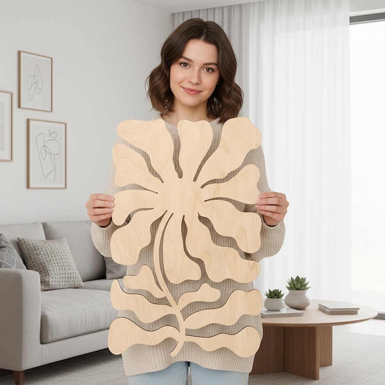 Peut inclure: Une grande d&eacute;coupe de fleur en bois de couleur claire. La fleur a plusieurs p&eacute;tales et une tige avec des feuilles. La d&eacute;coupe est tenue par une personne portant un pull de couleur claire, dans une pi&egrave;ce bien &eacute;clair&eacute;e avec un canap&eacute; et une table basse.
