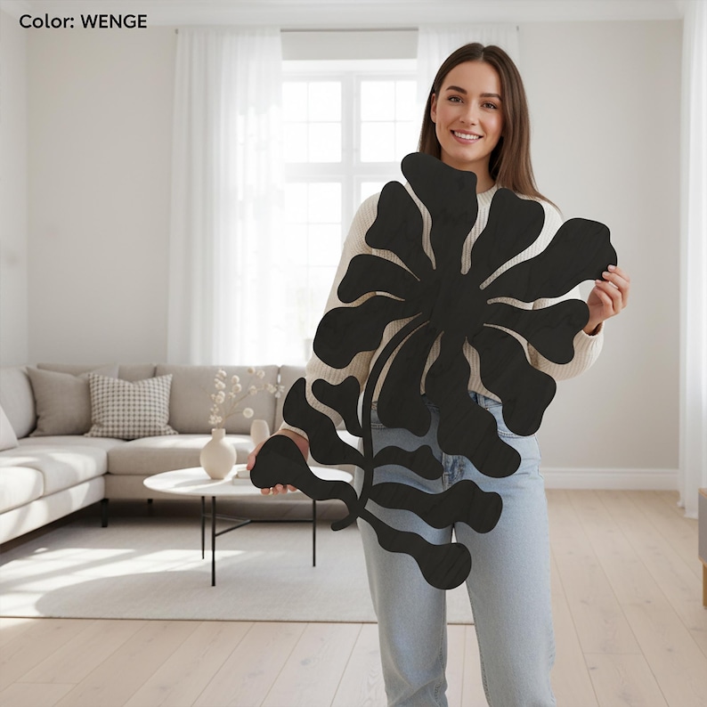 Peut inclure: Une grande &oelig;uvre d'art murale en bois noir avec un motif floral. L'&oelig;uvre est tenue par une personne portant un pull clair et un jean. Le texte "Color: WENGE" est dans le coin sup&eacute;rieur gauche.
