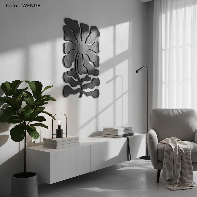 Peut inclure: Une &oelig;uvre d'art murale en bois noir en forme de fleur et de feuilles stylis&eacute;es. L'&oelig;uvre est fix&eacute;e sur un mur blanc. Le mot "WENGE" est dans le coin sup&eacute;rieur gauche. Un meuble blanc, une lampe et une plante en pot sont au premier plan.