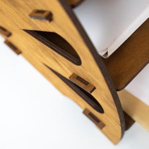 K&ouml;nnte beinhalten: Nahaufnahme eines Schreibtisch-Organizers aus Holz mit hellbraunem Finish. Der Organizer hat ein einzigartiges Design mit ausgeschnittenen Formen und dunkelbraunen Akzenten. Die Konstruktion umfasst kleine Holzd&uuml;bel zur Montage. Ein wei&szlig;es Objekt ist teilweise sichtbar.